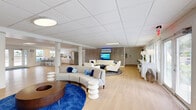 Located at 4314 Ginger Cove Drive in Tampa, FL, this single-story commercial space offers a generously sized, open-plan interior designed for both functionality and comfort. The floor plan is thoughtfully arranged into distinct zones while maintaining a seamless, flowing layout throughout.

The main area features light hardwood flooring, a drop-panel ceiling with recessed and flush-mounted lighting, and slender white support columns that define spaces without enclosing them. Multiple seating arrangements are distributed across the floor, including curved sofas, lounge chairs, and accent stools styled in neutral tones accented with vibrant blue — creating a cohesive coastal aesthetic. A textured stone accent wall serves as a focal point, anchoring a media area with a mounted flat-screen display.

Large windows and glass French doors line the perimeter, flooding the interior with natural light and providing views of the surrounding greenery. A dedicated kitchenette at one end includes a white island with bar seating, stainless steel appliances, and ample counter space. Adjacent to the kitchenette, a workstation area with desk space is well-suited for administrative use.

The overall ambiance is bright, modern, and welcoming, with a carefully curated coastal-inspired design that balances professional functionality with an inviting atmosphere.