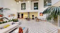 Located at 550 W Date St in San Diego's Little Italy neighborhood, this impressive lobby space makes an immediate statement through its bold architectural design and carefully curated interiors. The entrance greets visitors with a distinctive black-and-white patterned tile floor that anchors the expansive, open layout, while soaring double-height ceilings amplify the sense of grandeur and openness throughout.

A standout feature is the dramatic accent wall adorned with a grid of vibrant red acrylic panels suspended on vertical black rods, creating a striking artistic focal point visible from multiple vantage points. Large floor-to-ceiling windows flood the reception area with natural light, framing views of the surrounding San Diego streetscape. Rich warm-toned wood paneling and wood plank ceilings add texture and warmth, balancing the space's contemporary edge.

The lobby is divided into several distinct seating zones, each thoughtfully furnished with modern lounge chairs, sofas, and coffee tables. Curated artwork, decorative mirrors, and sculptural accents are displayed throughout, contributing to a gallery-like atmosphere. A glassed-in lounge area and a covered outdoor courtyard with lush greenery provide additional gathering spaces. The reception desk is seamlessly integrated, completing a welcoming, design-forward environment ideal for residents and visitors alike.