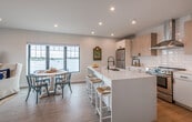 This thoughtfully designed single-floor residence showcases a bright, open-concept layout with a coastal aesthetic throughout. The heart of the home is a modern kitchen featuring a large white quartz island with an undermount sink, light wood upper cabinetry, crisp white lower cabinets with matte black hardware, a classic subway tile backsplash, stainless steel appliances, and a sleek range hood. The kitchen flows seamlessly into a generous living and dining area bathed in natural light from large windows and sliding glass doors that open to a balcony overlooking the water, offering sweeping waterfront views. Light hardwood flooring runs throughout, complementing the clean, neutral palette.

The living area provides a relaxed yet refined space, while the multiple bedrooms carry the coastal theme with natural textures and ample natural light. The primary bedroom includes a walk-in closet and a wall-mounted split system. A second bedroom features a large window framing waterfront scenery. The clean, modern bathroom is appointed with a soaking tub and shower combo, a floating vanity, and matte black fixtures. Recessed lighting throughout enhances the bright, airy ambiance of this well-appointed coastal retreat.