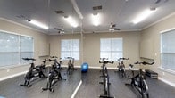 This single-floor fitness studio is purpose-built for indoor cycling and group exercise. The room is outfitted with multiple stationary spin bikes arranged to give each user ample space, making it well-suited for both individual training sessions and instructor-led cycling classes. A full-length mirrored wall spans one side of the room, enhancing the sense of openness while allowing participants to monitor their form during workouts. The flooring is a durable rubber gym surface, providing the grip and cushioning needed for active use. Two ceiling fans are mounted overhead, ensuring consistent air circulation throughout the room to keep the environment comfortable during intense exercise. Recessed fluorescent lighting provides bright, even illumination throughout the space. Natural light filters in through multiple windows, which also offer views of the surrounding greenery, adding a refreshing element to the workout atmosphere. Crown molding along the ceiling adds a subtle architectural refinement to the otherwise functional space. A stability ball is also present, indicating the room supports a variety of fitness activities beyond cycling. Overall, this studio combines practicality and comfort, creating an inviting environment for a focused fitness experience.