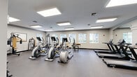 Located at 7350 NW Fourth St in Plantation, Florida, this fitness center offers a comprehensive, single-floor exercise facility designed to support a wide range of workout routines. The space is thoughtfully laid out with a generous open floor plan, providing ample room to move between equipment without feeling cramped.

The exercise room is outfitted with a robust selection of cardio machines, including multiple ellipticals and treadmills from True Fitness, as well as a recumbent bike. Strength training is well represented through a cable/multi-function machine, free weights and a dumbbell rack, medicine balls, stability balls, and a weight bench. A wall-mounted flat-screen TV keeps members entertained during workouts.

Overhead, large recessed panel lighting fixtures provide bright, even illumination throughout the space. Multiple windows along the walls invite natural light and offer views of the surrounding outdoor greenery and neighboring buildings, creating an open, airy atmosphere. Full-length mirrors on one wall enhance the sense of space and allow for form-checking during exercise.

The durable dark rubber flooring offers both safety and comfort underfoot. The neutral gray and white color palette gives the room a clean, professional aesthetic. Motivational signage adds an energetic touch, completing a functional and welcoming environment suited for daily fitness use.