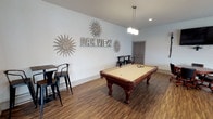 The Oak View amenity space is a thoughtfully designed, single-floor community hub that blends recreation and relaxation into one cohesive environment. The game room serves as a focal point, centered around a full-size pool table with a warm cherry wood finish and cream felt surface. A shuffleboard table adds to the entertainment options, while black metal bar-height tables and chairs provide casual seating throughout. Decorative sunburst wall art and bold "Oak View" marquee lettering lend the space a distinct identity. Pendant lighting fixtures with frosted glass shades illuminate the area with warm, inviting light.

A row of large windows along one wall floods part of the space with natural light and offers views of the surrounding community. The game room opens seamlessly into a spacious lounge area, which features soaring vaulted ceilings with exposed beam detailing and a statement ceiling fan. This lounge is furnished with red accent chairs, a dark leather sectional, and a central dining-height table set atop a patterned area rug over large-format tile flooring. A dedicated workstation with a desk and computer equipment provides a functional corner for residents. Rich wood-look flooring throughout the game room ties the entire space together with a warm, polished finish.