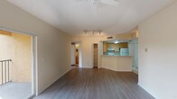 This single-floor residence features a thoughtfully designed open-concept layout with consistent wood-style flooring flowing throughout the main living areas. The spacious living room is well-lit by natural light from a sliding glass door that opens to a private balcony with outdoor views, complemented by a ceiling fan and track lighting overhead. A textured ceiling adds visual interest throughout the space.

The kitchen is positioned adjacent to the living area and features a peninsula-style breakfast bar, light wood cabinetry extending to the ceiling, a double sink, white appliances including an electric range and refrigerator, and tile flooring. A louvered closet door provides additional pantry or storage access.

A dedicated laundry area with stacked units is conveniently located off the main living space. The bedroom offers a bright, airy feel with a large window that brings in ample natural light. The full bathroom includes a single-sink vanity with light wood cabinetry, a tub-shower combination, and tile flooring. The overall color palette of soft neutrals creates a clean, cohesive ambiance throughout the unit.