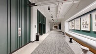 This single-floor commercial space in St. Louis, Missouri, delivers a refined blend of industrial and contemporary design throughout its expansive layout. The main corridor is a standout feature, stretching at length with a textured gray carpet runner anchored by polished concrete flooring on either side. Exposed overhead infrastructure—including white conduit, track lighting, and pendant fixtures—contrasts with clean white walls, while deep green accent panels frame doorways and restroom entries. Framed artwork lines one corridor wall above a continuous wood bench with sculptural stool accents, contributing an art-gallery sensibility to the circulation space.

A dramatic glazed wall opens onto an interior courtyard atrium, offering views of lush plantings and communal seating beyond steel-framed glass. A wood-platform seating area with a slatted bench faces this transparent boundary, creating a calm transitional zone. The lounge area features a latticed wood ceiling, backlit built-in shelving, textured accent walls, and a curated furniture arrangement centered on a round coffee table.

Restrooms are finished with classic black-and-white hexagonal tile flooring, subway-style wall tile, and bold black partition frames with cityscape-patterned dividers. A dedicated bike storage room with wall-mounted racks and polished concrete flooring rounds out the amenities.