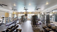 Located at 4401 E 46th St in North Little Rock, AR, this fitness center offers a generously sized, single-floor workout space designed to accommodate a full range of exercise needs. The open floor plan is covered in durable speckled rubber flooring, providing both safety and comfort underfoot throughout the entire area.

The facility is outfitted with an extensive array of strength training equipment, including multiple cable and selectorized weight machines, adjustable benches, and a well-stocked dumbbell rack ranging across a wide variety of weights. Full-length wall mirrors line key sections of the room, amplifying the sense of space and allowing users to monitor their form during workouts.

Natural light streams in through numerous large windows along the perimeter walls, creating a bright and energizing atmosphere. Multiple ceiling fans are distributed throughout the space, ensuring consistent air circulation and comfort during intense training sessions. Recessed lighting supplements the natural light for well-lit conditions at all times.

Motivational signage adorns the walls, reinforcing an encouraging workout environment. The warm-toned walls and crown molding details lend a polished, finished aesthetic to the space. Views through the windows suggest proximity to an outdoor pool area, adding to the overall appeal of this comprehensive fitness facility.