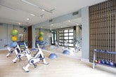Located at 2204 Duluth Highway 120 in Duluth, Georgia, this single-floor fitness studio is thoughtfully designed to deliver a bright, open, and motivating workout environment. The space is defined by its standout architectural feature: full-panel glass garage doors framed in sleek black metal that span a significant portion of the wall, allowing abundant natural light to pour in and offering unobstructed views of the surrounding lush green landscaping. When opened, these doors seamlessly connect the interior workout space to the outdoors, creating an airy, open-air training experience.

The light wood flooring throughout lends a clean, contemporary aesthetic that complements the cool gray-blue walls and recessed ceiling lighting. The studio is equipped with a row of True-brand stationary spin bikes, positioned to take advantage of the outdoor views. Along the opposite end of the room, a well-organized equipment station houses stability balls in multiple sizes, resistance bands, medicine balls, and a BOSU balance trainer, catering to a variety of fitness disciplines. A wall-mounted fan provides additional airflow, and a full-length mirror enhances the sense of spaciousness. The overall ambiance is modern, functional, and energizing.