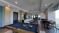 This contemporary amenity lounge is a thoughtfully designed, single-floor space that seamlessly blends recreation, entertainment, and social gathering areas into one cohesive environment. At the forefront is a full-size billiards table with a rich wood frame and navy felt surface, setting a polished tone for the recreation zone. Beyond it, a well-appointed kitchen anchors the space, featuring dark cabinetry, stainless steel appliances, and a striking stone tile accent wall. A large white island with pendant lighting overhead serves as a natural gathering point, offering bar-style seating for casual dining or conversation. The flooring transitions from tile near the kitchen to warm wood-toned planks throughout, adding visual depth and flow. Further into the space, a cozy fireplace lounge area provides a more relaxed setting, complemented by comfortable seating and a wall-mounted television. Multiple dining tables with high-back chairs are distributed throughout, accommodating both intimate and group gatherings. Recessed lighting runs across the ceiling alongside in-ceiling speakers, ensuring the space is well-lit and acoustically equipped. Large windows allow natural light to filter in, enhancing the modern, upscale ambiance of this versatile communal space.