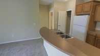 This two-floor space is designed with a practical, open-concept layout that connects the kitchen and living areas seamlessly. The kitchen features a curved peninsula with a laminate countertop and a double stainless steel sink, providing ample workspace and a natural dividing point between the kitchen and the adjoining carpeted living area. Light-toned cabinetry is visible near the refrigerator, offering functional storage. The walls are finished in a neutral warm beige, contributing to a clean and cohesive aesthetic throughout.

The living area is carpeted in a neutral tone, complementing the overall color palette and adding a comfortable feel underfoot. Two doorways are visible from the main living space — one leading to what appears to be a closeted storage area, and another opening to an additional closet, suggesting well-considered storage solutions. The ceilings are of a standard height, and the overall layout feels open and uncluttered. White trim and baseboards provide clean contrast against the walls and carpet. The two-floor configuration of this space suggests additional living or sleeping areas on a separate level, expanding its overall functionality.