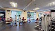 This fitness center offers a spacious, single-floor layout designed to accommodate a full range of workout needs. The space is divided into distinct functional zones, seamlessly organized for an efficient exercise experience. The cardio area features a row of elliptical trainers and treadmills, all positioned near large windows that flood the space with natural light and offer views of the surrounding greenery outside. The strength training section is well-stocked with a variety of cable and selectorized machines, including leg press, shoulder, and chest equipment, complemented by a dedicated free weights area with a dumbbell rack visible along the wall. Large wall-to-wall mirrors line one side of the facility, visually expanding the space and allowing users to monitor their form during workouts. The flooring throughout is a dark, durable rubber material, ideal for heavy equipment and high-traffic use. Overhead, a drop tile ceiling with recessed fluorescent lighting ensures the space is consistently and evenly illuminated. Accents of deep red on the walls provide a bold, energetic atmosphere, while structural columns punctuate the open floor plan. The overall ambiance is clean, functional, and professional, making it a comprehensive facility suited for a wide variety of fitness routines.