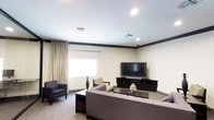 This single-floor space offers a welcoming and functional layout centered around a thoughtfully designed lounge area. The main sitting room is furnished with a large sectional sofa in a neutral gray tone, complemented by accent chairs — including a patterned armchair and a mustard-yellow side chair — arranged to encourage conversation and relaxed viewing. A dark wood media console anchors one wall, supporting a large flat-screen television, while a low-profile coffee table sits at the center of the seating arrangement, decorated with a ceramic accent piece that adds a touch of warmth.

The room features recessed lighting set into smooth, light-toned ceilings with dark crown molding that provides a clean, defined contrast along the upper perimeter. A window dressed with layered curtains — a sheer roller shade beneath warm beige drape panels — allows for adjustable natural light while maintaining privacy. The neutral wall tones and carpeted flooring contribute to the overall warm, understated ambiance of the space. The layout is open and proportioned generously, making it well-suited for communal relaxation or informal gatherings. Overall, the space strikes a balance between comfort and practicality with a cohesive, neutral color palette throughout.