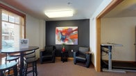 This well-appointed single-floor space is designed to foster comfort and community. The entry vestibule makes a strong first impression, featuring large glass doors that flood the area with natural light, warm oak wood trim, and clean tile flooring with a welcoming mat. From the entry, the space opens into a corridor accented with elegant wall sconces, sage green upholstered armchairs, and a decorative gold side table, creating an inviting transitional area.

The lounge area is a standout feature, characterized by a dramatic dark accent wall serving as a backdrop for a large, vibrant abstract painting bursting with bold reds, oranges, and blues. Two sleek black leather armchairs flank a small side table, complemented by a high-top table and bar stools positioned near a window offering natural light. The carpeted flooring adds warmth and acoustic comfort throughout.

An adjacent gathering or activity room continues the dark accent wall theme, featuring a ceiling fan with light fixture, a high-top table with stools, a decorative faux tree, and a leaning bookshelf for literature or resources. A corkboard provides a functional touch for community notices. Together, these spaces offer a cohesive, thoughtfully designed environment ideal for relaxing, socializing, or casual meetings.