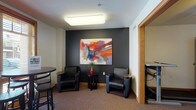 This well-appointed single-floor space is designed to foster comfort and community. The entry vestibule makes a strong first impression, featuring large glass doors that flood the area with natural light, warm oak wood trim, and clean tile flooring with a welcoming mat. From the entry, the space opens into a corridor accented with elegant wall sconces, sage green upholstered armchairs, and a decorative gold side table, creating an inviting transitional area.

The lounge area is a standout feature, characterized by a dramatic dark accent wall serving as a backdrop for a large, vibrant abstract painting bursting with bold reds, oranges, and blues. Two sleek black leather armchairs flank a small side table, complemented by a high-top table and bar stools positioned near a window offering natural light. The carpeted flooring adds warmth and acoustic comfort throughout.

An adjacent gathering or activity room continues the dark accent wall theme, featuring a ceiling fan with light fixture, a high-top table with stools, a decorative faux tree, and a leaning bookshelf for literature or resources. A corkboard provides a functional touch for community notices. Together, these spaces offer a cohesive, thoughtfully designed environment ideal for relaxing, socializing, or casual meetings.