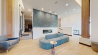 This thoughtfully designed single-floor communal space blends contemporary architecture with comfortable, functional interiors. The central lounge anchors the layout, highlighted by a dramatic floor-to-ceiling accent wall clad in a teal chevron-patterned tile, behind which a sleek linear fireplace serves as a striking focal point. Light wood flooring runs throughout, complementing the crisp white walls and contributing to an open, airy atmosphere. A sculptural teal curved sofa anchors the seating area, paired with accent chairs and minimalist side tables.

Floor-to-ceiling windows and glass-paneled walls flood the entry and lounge areas with natural light, framing outdoor views and reinforcing a strong indoor-outdoor connection. Wood accent columns add warmth to the otherwise modern palette.

A dedicated media lounge features a built-in bench with dual wall-mounted televisions set against a deep purple textured tile backdrop, complemented by a mix of gray and orange upholstered ottomans and soft patterned carpeting. An adjacent seating nook offers a large sectional sofa and curated wall art for a more intimate setting.

The kitchenette is outfitted with pale blue cabinetry, black hardware, a penny tile backsplash, and full-size appliances. A decorative chain curtain subtly partitions this area from the lounge, adding a refined, design-forward detail throughout.