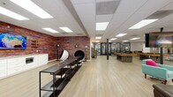Located at 919 N Stone Ave in Tucson, Arizona, this single-floor community amenity space is designed to offer residents a dynamic and engaging environment for relaxation, recreation, and fitness. The interior is anchored by light wood-look flooring throughout, complemented by a drop ceiling with recessed panel lighting that keeps the atmosphere bright and welcoming.

The lounge area features large windows that invite natural light and offer views of the outdoor pool and surrounding greenery. Comfortable seating, including teal accent chairs, a large sectional sofa, and eclectic side tables, creates an inviting gathering space accented by vibrant Southwestern-themed artwork.

Adjacent to the lounge, the game room stands out with a bold, multicolored textured accent wall serving as a striking visual focal point. This area includes multiple pool tables, a wall-mounted television, a built-in kitchenette with white cabinetry and a beverage cooler, shelving units, and unique pod-style lounge chairs that add a playful, modern touch.

The fitness center is enclosed by sleek floor-to-ceiling glass walls and doors, providing full visibility into the well-equipped space featuring treadmills, cycling machines, and functional training equipment. Together, these amenities create a cohesive, energetic community hub suited for socializing and active living.