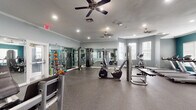 This fitness center offers a well-organized, single-level exercise space designed to accommodate a range of workout routines. The room is finished with durable rubber flooring throughout, providing a practical and cushioned surface suitable for both cardio and strength training activities. Recessed lighting and multiple ceiling fans are installed overhead, ensuring the space remains well-lit and properly ventilated during use.

The main workout area features an assortment of strength training machines, including cable systems, a leg press, and a chest press, along with free weights and a colorful dumbbell rack. Cardio equipment, including treadmills, an elliptical, and a stationary bike, is distributed throughout the space. Full-length mirrors line one wall, visually expanding the room and allowing users to monitor their form during exercise.

A secondary area, distinguished by its bold teal accent wall, connects to the main workout floor and houses additional cardio machines, including treadmills. This section provides direct access via glass-paneled doors to an adjacent corridor. Natural light enters through multiple windows fitted with blinds, contributing to an open and energizing atmosphere. Crown molding details add a refined architectural touch throughout the space.