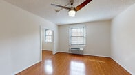 This single-floor unit offers a well-organized layout with a range of functional and comfortable living spaces. The main living and bedroom areas are finished with warm-toned hardwood flooring that reflects natural light, creating an inviting atmosphere throughout. Ceiling fans are present in the primary rooms, providing both ventilation and a classic aesthetic complement to the textured ceilings. Multiple windows fitted with horizontal blinds allow ample natural light to fill the rooms, enhancing the open and airy feel of the space.

The kitchen is efficiently designed in a galley-style configuration, featuring warm wood upper and lower cabinetry that provides generous storage. Dark countertops contrast neatly with white appliances, including an electric range, over-the-range microwave, and refrigerator. The kitchen floor is finished in neutral tile, clearly delineating it from the hardwood in the adjoining areas. A window at the end of the kitchen brings in additional natural light.

The bedroom is a well-proportioned room with hardwood floors, a ceiling fan, a wall-mounted HVAC unit, and access to a closet. The bathroom is clean and bright, finished in white tile with a full bathtub, toilet, and vanity, and features a window that allows for natural ventilation. Overall, the space is practical and well-lit with a cohesive, neutral design throughout.