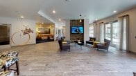 Located at 530 11th Ave N in Nashville, Tennessee, this single-floor communal amenity space is thoughtfully designed to encourage social interaction and relaxation. The layout flows seamlessly across multiple distinct zones, each with its own character while maintaining a cohesive contemporary aesthetic throughout.

The main lounge area features natural stone-look tile flooring and serves as the central hub of the space, furnished with sleek charcoal armchairs accented with mustard yellow cushions, arranged around a textured stone accent wall housing a mounted flat-screen television. Pendant lighting and sheer-curtained windows invite natural light from the exterior.

A dedicated lounge nook is carpeted in deep navy blue and anchored by a large cognac leather sectional sofa, accented with chartreuse throw pillows. A geometric sculptural coffee table sits at its center, while curated contemporary artwork adorns the walls, creating an intimate, gallery-like atmosphere.

A game room area features wide-plank wood-look flooring, a large-scale wall-mounted Scrabble board, casual seating, and an oversized garage-style door that opens directly to an outdoor patio, blurring the boundary between indoor and outdoor spaces.

The kitchenette is outfitted with quartz countertops, dark wood cabinetry, stainless steel appliances, a sink, and globe pendant lighting. A cozy dining nook with banquette seating and a colorful accent wall completes the space.