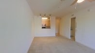 This single-floor residence offers a thoughtfully designed layout that balances openness with functional living. The main living area is finished with neutral-toned wall-to-wall carpeting, creating a warm and inviting atmosphere. Bright white walls reflect natural and artificial light throughout, contributing to an airy, spacious feel.

A defining feature of this space is the pass-through kitchen window, which connects the kitchen to the main living area, promoting an open and sociable flow between spaces. The kitchen is equipped with warm wood-toned cabinetry that adds a natural, grounded element to the otherwise light and neutral palette. Built-in appliances, including a microwave and stovetop range, are integrated seamlessly within the cabinetry. A track lighting system mounted on the ceiling above the pass-through provides focused illumination across both the kitchen and living areas.

Additional doorways visible from the main living space suggest further rooms beyond, hinting at a layout with multiple functional zones. Overhead HVAC vents indicate a centralized climate control system throughout. The overall ambiance of this space is clean, minimal, and move-in ready, offering a versatile and well-lit interior suited to a variety of living arrangements.