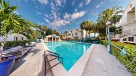 This outdoor amenity space centers around a generously sized swimming pool with crystal-clear turquoise water, set within a well-maintained deck area that accommodates multiple lounge chairs positioned for sunbathing. The pool features entry steps with metal handrails, making it accessible and functional for everyday use.

The surrounding environment is distinctly tropical in character, with tall palm trees and dense green hedging framing the perimeter and providing natural privacy. A covered cabana or pavilion structure is visible at one end of the deck, equipped with additional seating, offering a shaded retreat from the sun. Vibrant blue umbrellas and lounge chairs add a cohesive, resort-like aesthetic to the space.

The multi-story building backdrop is finished in a light neutral exterior, contributing a clean, coastal architectural character to the setting. The open sky above, often bright with scattered clouds, enhances the airy, sun-drenched atmosphere. Decorative lighting fixtures are present along the pool surround, suggesting the space is also functional and inviting during evening hours. Overall, the area presents a relaxed, well-appointed outdoor retreat with a distinctly warm-weather, leisure-focused ambiance.