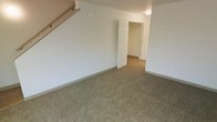 This two-floor residential space offers a functional and straightforward layout designed for comfortable everyday living. The image reveals a carpeted room that serves as a primary living or bedroom area on one of the floors, featuring neutral-toned wall-to-wall carpeting that adds warmth underfoot. The walls are finished in a clean, light gray-white tone, contributing to a bright and airy feel throughout the space.

A staircase is visible along one wall, with a handrail extending diagonally, indicating direct interior access between the two floors. The open doorway visible from this room leads into an adjacent area, where a closet with a paneled door can be seen, suggesting practical built-in storage. A second paneled door is also visible nearby, hinting at additional rooms or storage beyond.

The ceiling-mounted lighting provides consistent illumination, keeping the space well-lit. The overall design is clean and understated, with simple architectural details such as white panel doors and standard baseboards throughout. The layout appears to allow for easy flow between rooms, making the space adaptable for various furniture arrangements and uses across both levels.
