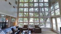 This single-floor space makes an immediate impression with its most defining architectural feature: a towering, grid-paned window wall that spans the full height and width of the main living area. The expansive glazing allows an abundance of natural light to pour into the interior throughout the day, creating a bright and airy atmosphere that feels seamlessly connected to the lush outdoor surroundings. Mature trees visible through the glass provide a verdant, ever-changing backdrop that enhances the sense of calm and openness.

The soaring ceiling height amplifies the grandeur of the space, lending it a sense of volume and scale well beyond what its single-floor layout might suggest. The living area is thoughtfully appointed with a neutral color palette — warm beige tones on the walls and flooring complement the natural wood and upholstered furnishings, including a leather sofa, cushioned armchairs, and a dark-finish coffee table. Subtle accent lighting from table lamps adds warmth to balance the abundant daylight.

Overall, the space strikes a balance between architectural drama and comfortable livability, making it well-suited for both relaxed everyday use and entertaining. The interplay of light, greenery, and refined finishes gives the space a welcoming yet sophisticated ambiance.