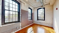 This thoughtfully converted loft-style unit blends historic industrial character with modern finishes across a compact yet efficient single-floor layout. The open living and kitchen area serves as the heart of the space, showcasing original whitewashed brick walls, warm hardwood flooring, and soaring ceilings with exposed ductwork and wooden planking that nod to the building's industrial past. Large, arched black-framed multi-pane windows flood the space with natural light and frame outdoor views, reinforcing the unit's converted warehouse aesthetic. The kitchen features white cabinetry, stainless steel appliances, and a clean, functional layout integrated seamlessly into the main living area.

Both bedrooms continue the industrial aesthetic, with painted brick accent walls and generously sized arched windows. Soft carpet flooring provides comfort underfoot, and each room includes a closet for storage. The primary bedroom features notably tall ceilings with exposed ductwork overhead.

The bathroom offers a clean, contemporary finish with a granite-topped white vanity, a framed mirror, and light tile flooring. An in-unit stacked washer and dryer is conveniently accessible nearby. A hallway connects the private areas of the unit, completing this character-rich, efficiently designed space.