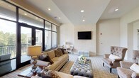Located at 260 Leigh Farm Rd in Durham, NC, this refined single-floor lounge space is designed with a sophisticated aesthetic that balances comfort and elegance. The main living area is anchored by a striking textured wood-plank accent wall that rises to an angled ceiling, adding depth and warmth to the neutral color palette throughout. Recessed lighting illuminates the space evenly, highlighting plush seating arrangements that include tufted wingback chairs, a classic rolled-arm sofa, and a tufted silver ottoman serving as a central focal point. A second seating zone is centered around a wall-mounted flat-screen television set against a light stone-textured feature wall, creating an inviting media area. Large floor-to-ceiling windows and glass doors flood the interior with abundant natural light while offering direct access to a private outdoor balcony. The balcony is outfitted with all-weather wicker lounge furniture and a glass coffee table, providing an open-air retreat with views of the surrounding community. Patterned area rugs define each seating zone, while curated artwork and decorative accents contribute to the space's polished, upscale character. The overall ambiance is warm, stylish, and thoughtfully appointed throughout.