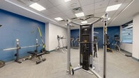 Located at 104 Washington St in Dover, NH, this fitness center offers a thoughtfully designed exercise space that accommodates a comprehensive range of workout activities. The room features a clean, modern aesthetic with a combination of bold blue accent walls and crisp white finishes, complemented by herringbone-patterned commercial carpet tile flooring throughout.

The space is well-lit by a recessed drop-panel ceiling with evenly distributed flush lighting, creating a bright and energizing atmosphere. A standout architectural feature is the large glass partition wall with frosted detailing, which allows natural light to filter in while maintaining a sense of openness and connectivity to adjacent areas.

The cardio zone is lined with multiple treadmills and elliptical machines, providing ample options for aerobic exercise. A dedicated strength training area includes a Hoist cable machine system, free weights, a weight bench, and additional resistance equipment, offering a well-rounded selection for full-body workouts. Cycling equipment and TRX-style suspension trainers round out the versatile equipment offering.

Additional amenities include a water fountain station, a fire safety cabinet, and seating near the entry. The layout flows efficiently, allowing users to move freely between equipment zones. This facility is well-suited for daily fitness routines in a professional and polished environment.