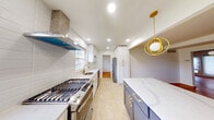 This two-floor home thoughtfully combines mid-century architectural character with contemporary renovations throughout. The main living area immediately draws attention with its distinctive white-painted plank ceiling accented by rich, dark wood exposed beams, creating a striking overhead focal point. Warm parquet hardwood flooring in a classic block pattern runs throughout the living spaces, adding texture and visual depth underfoot. A central fireplace with a raised hearth anchors the main living room, flanked by built-in shelving, while large windows fitted with roller shades invite natural light into the generously proportioned space.

The kitchen has been fully renovated to a high standard, featuring white shaker cabinetry with brushed gold hardware, contrasted elegantly by a navy blue island with a quartz waterfall countertop. A professional-grade stainless steel gas range sits beneath a sleek stainless hood, complemented by subway tile backsplash, a gold-toned faucet, and a stainless refrigerator. Recessed lighting and a sculptural gold pendant light complete the refined aesthetic.

The bathrooms reflect careful attention to detail, with one featuring floor-to-ceiling grey tile, a glass-enclosed marble shower, and gold accent hardware. Bedrooms are bright and well-proportioned, continuing the parquet flooring, with ample closet space and multiple windows offering natural light.