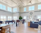 Located at 4800 Tanner St in Gillette, Wyoming, this 2,920 square foot single-floor clubhouse is a thoughtfully designed community gathering space that combines modern aesthetics with functional versatility. The open-concept layout is anchored by dramatically soaring ceilings adorned with exposed industrial-style ductwork, elegant chandelier lighting, and ceiling fans that enhance the airy, loft-like atmosphere throughout.

The main living area features multiple seating arrangements, including plush blue upholstered sofas and lounge chairs centered around a floor-to-ceiling stone fireplace with a wall-mounted television above — creating an inviting focal point for socializing. Light oak hardwood flooring runs throughout, complementing the crisp white walls and abundant natural light streaming in from expansive upper clerestory windows and large ground-level windows that frame sweeping views of the surrounding Wyoming landscape.

A dedicated dining zone offers round and rectangular tables with upholstered chairs, while bar-height tables provide additional casual seating options. Western-themed equestrian artwork adorns the walls, adding a regional character to the space. A pool table, arcade game stations, and a built-in bookcase further enhance the recreational offerings.

Additional amenities include a drinking fountain station set against a decorative accent wall, and glass double doors providing easy exterior access. The clubhouse offers a welcoming, multi-purpose environment ideal for community events and gatherings.