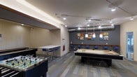 This single-floor recreational space is thoughtfully designed as a social hub for entertainment and leisure. The room features a contemporary aesthetic, with a combination of warm neutral tones and bold accent walls in deep gray, creating an energetic yet inviting atmosphere. Stylish pendant lighting hangs above a full-size pool table, which serves as the centerpiece of the room, while track lighting throughout ensures the space is well-lit. A foosball table is positioned near the entrance, adding to the variety of gaming options available. Along one wall, a bar-height counter with blue upholstered stools provides a dedicated seating area ideal for spectators and socializing. Colorful wall art and graphic prints add personality and visual interest to the space. The flooring features a mixed wood-tone plank design that complements the modern industrial aesthetic, with exposed ductwork on the ceiling adding to the urban character of the room. Cue sticks are conveniently mounted on the wall near the pool table, and subtle cove lighting along the ceiling perimeter adds a polished, refined finish. Overall, this game room delivers a well-appointed and versatile environment suited for casual gatherings and recreational activities.