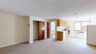 Located at 9 Chester St in Cambridge, MA, this single-floor unit offers a practical and straightforward layout well-suited for everyday living. The open main living area is finished with wall-to-wall carpeting and neutral cream-toned walls, creating a clean, bright atmosphere with ample room to arrange furniture as desired. The kitchen is integrated into the main space and is equipped with warm-toned wood cabinetry, a tile backsplash, white countertops, a double-basin sink, an electric range with oven, a dishwasher, and a refrigerator. A window in the kitchen area allows natural light to filter in, and a wall-mounted air conditioning unit provides climate control. The main living area also features a closet with a hanging rod, offering convenient storage. The bathroom includes a full-size soaking tub and shower combination surrounded by clean white subway tile, with a window above providing natural light and ventilation. A separate toilet and sink area complete the bathroom space. Baseboard heating is present throughout, ensuring warmth during Cambridge's colder months. The overall design is functional and uncluttered, making this a practical unit in a well-established Cambridge neighborhood.