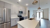 This single-floor space showcases a thoughtfully designed, open-concept kitchen that serves as a central highlight. The kitchen features soft gray shaker-style cabinetry extending to the upper walls, paired with crisp white quartz countertops that provide ample prep and workspace. A generously sized island anchors the layout, complete with an undermount sink and a brushed nickel pull-down faucet, making it both a functional and social focal point. Stainless steel appliances, including an electric range and an over-the-range microwave, lend a polished, contemporary feel. A classic white subway tile backsplash adds texture and visual interest behind the cooking area. Warm-toned wood-look flooring runs throughout, complementing the neutral color palette. Recessed lighting illuminates the space efficiently, while brushed gold pendant lights above the island introduce an elegant accent. A window visible in the adjoining living area allows natural light to filter into the open layout, enhancing the overall brightness and airy ambiance of the space.