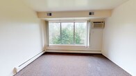 This one-bedroom, one-bathroom unit in Westland, Michigan offers a practical and comfortable layout across a single floor. The spacious living room is carpeted and features a large picture window that frames lush, tree-lined views, filling the room with natural light. A wall-mounted air conditioning unit and baseboard heating ensure year-round comfort. The bedroom is generously sized with plush carpeting, a ceiling fan with light fixture, a window overlooking greenery, and a closet with built-in shelving for ample storage. An additional closet is accessible from the main living area. The kitchen is efficiently designed with light wood-look vinyl flooring, white cabinetry, a full-size refrigerator, an electric range with overhead ventilation, and a dedicated pantry or storage closet nearby. The bathroom features white tile flooring, a full bathtub with classic blue tile surround, a toilet, and a wood-toned vanity with a white countertop. Baseboard heating is present throughout the unit, and the overall color palette of neutral whites and beiges creates a clean, bright atmosphere. The treetop views visible from multiple rooms add a serene, natural quality to this well-maintained Westland residence.