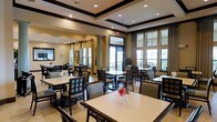 This sophisticated dining space is designed to offer a refined yet welcoming environment for guests. The room is anchored by a striking coffered ceiling with dark wood trim detailing, adding architectural depth and a sense of grandeur to the interior. Recessed lighting complements the natural light that pours in through a series of large, floor-to-ceiling windows along the exterior wall, dressed with elegant drapery panels that frame the views of the surrounding greenery outside.

The dining area is generously appointed with multiple tables, each set with light-toned surfaces and accompanied by dark wood-framed chairs with upholstered seats, striking a classic balance between style and comfort. Fresh floral accents on the tables add a cheerful, welcoming touch to the overall ambiance.

Structural columns punctuate the space, reinforcing the room's traditional architectural character, while light-toned flooring throughout creates a clean, airy foundation. A secondary dining or buffet area is visible in the background, suggesting the space is well-suited to accommodate a range of dining occasions. The warm neutral color palette, combined with the thoughtful layout and abundant light, makes this dining room both functional and aesthetically inviting.
