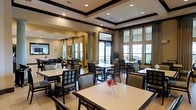 This sophisticated dining space is designed to offer a refined yet welcoming environment for guests. The room is anchored by a striking coffered ceiling with dark wood trim detailing, adding architectural depth and a sense of grandeur to the interior. Recessed lighting complements the natural light that pours in through a series of large, floor-to-ceiling windows along the exterior wall, dressed with elegant drapery panels that frame the views of the surrounding greenery outside.

The dining area is generously appointed with multiple tables, each set with light-toned surfaces and accompanied by dark wood-framed chairs with upholstered seats, striking a classic balance between style and comfort. Fresh floral accents on the tables add a cheerful, welcoming touch to the overall ambiance.

Structural columns punctuate the space, reinforcing the room's traditional architectural character, while light-toned flooring throughout creates a clean, airy foundation. A secondary dining or buffet area is visible in the background, suggesting the space is well-suited to accommodate a range of dining occasions. The warm neutral color palette, combined with the thoughtful layout and abundant light, makes this dining room both functional and aesthetically inviting.