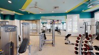 Located at 4000 Coleway Dr in Holly Springs, NC, this single-floor fitness center is a thoughtfully designed facility that combines energetic aesthetics with a broad range of exercise equipment. The space is immediately distinguished by its bold teal and yellow color scheme, which carries across the walls and ceiling accents, creating a lively and motivating atmosphere throughout.

The main workout floor is generously proportioned, housing an extensive selection of strength training machines, free weights, a rowing machine, and additional cable and resistance equipment. Large floor-to-ceiling mirrors line the walls, amplifying the sense of space, while mounted flat-screen TVs provide entertainment during workouts. Recessed lighting and ceiling fans ensure a comfortable environment.

A dedicated cardio zone features a row of treadmills and elliptical machines, all positioned beneath large, white-shuttered windows that invite abundant natural light and offer views of the surrounding community.

A separate studio room with warm hardwood flooring is outfitted with spin bikes, resistance bands, and free weights, making it well-suited for cycling sessions and group fitness classes. This room also benefits from wall-to-wall mirrors and a mounted display screen, supporting both guided and independent workouts.

Throughout the facility, natural light flows in generously from multiple windows, enhancing the open, airy feel of the space and creating an inviting environment for all fitness levels.