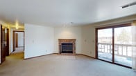 This single-floor space centers around a welcoming living area that combines functional design with comfortable, understated details. The room is finished with neutral-toned carpet that extends throughout, contributing to a warm and cohesive feel underfoot. Crisp white walls and a smooth ceiling provide a clean, bright backdrop, while recessed lighting and a ceiling vent ensure practical climate and lighting control.

The focal point of the living area is a classic brick fireplace, featuring a wooden mantel and glass-paneled doors that add both character and charm. A raised brick hearth extends the fireplace's visual presence and grounds the room. Adjacent to the fireplace, a wood-framed sliding glass door provides direct access to an outdoor balcony, allowing natural light to filter into the space and offering a view of the exterior surroundings.

Warm wood-toned baseboards run along the lower edges of the walls, adding a subtle contrast to the lighter finishes throughout. The overall layout is open and versatile, providing ample room for a variety of furniture arrangements. The space presents a practical yet inviting atmosphere suited to everyday living.