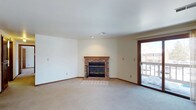 This single-floor space centers around a welcoming living area that combines functional design with comfortable, understated details. The room is finished with neutral-toned carpet that extends throughout, contributing to a warm and cohesive feel underfoot. Crisp white walls and a smooth ceiling provide a clean, bright backdrop, while recessed lighting and a ceiling vent ensure practical climate and lighting control.

The focal point of the living area is a classic brick fireplace, featuring a wooden mantel and glass-paneled doors that add both character and charm. A raised brick hearth extends the fireplace's visual presence and grounds the room. Adjacent to the fireplace, a wood-framed sliding glass door provides direct access to an outdoor balcony, allowing natural light to filter into the space and offering a view of the exterior surroundings.

Warm wood-toned baseboards run along the lower edges of the walls, adding a subtle contrast to the lighter finishes throughout. The overall layout is open and versatile, providing ample room for a variety of furniture arrangements. The space presents a practical yet inviting atmosphere suited to everyday living.