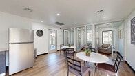 Located at 8330 N 19th Ave in Phoenix, Arizona, this single-floor community or amenity space offers a clean, modern design tailored for casual dining and social gatherings. The interior is finished with warm-toned, wide-plank wood flooring that runs throughout, complemented by crisp white walls and recessed lighting that keeps the space bright and welcoming.

The dining area serves as the central feature, accommodating multiple round white tables paired with contemporary dark-framed chairs. A wall-mounted flat-screen television is positioned above one seating area, adding a functional entertainment element. Decorative touches such as framed artwork, potted plants, and small centerpieces contribute to an inviting atmosphere.

A standout architectural element is the row of sleek, floor-to-ceiling glass-paneled partition walls with slim aluminum frames, creating semi-private lounge alcoves furnished with comfortable seating. These transparent dividers maintain an open, airy feel while offering flexible use of the space.

Along one wall, a compact kitchenette includes dark cabinetry, a stainless steel refrigerator, and counter space for basic food and beverage needs. A glazed exterior door provides natural light and direct access to an outdoor area. The overall design balances functionality with a contemporary aesthetic, making it a versatile and well-appointed shared space.