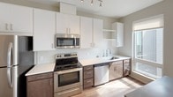 This thoughtfully designed single-floor unit offers a cohesive and modern aesthetic throughout. The kitchen is a standout feature, showcasing a galley-style layout with sleek white upper cabinetry and contrasting dark lower cabinets, white quartz countertops, a classic white tile backsplash, and a full suite of stainless steel appliances including an electric range, built-in microwave, dishwasher, and refrigerator. Track lighting overhead illuminates the workspace efficiently. Warm wood-look flooring flows consistently throughout the unit, connecting each area with a sense of continuity and warmth. The living area is open and filled with natural light from a large window that frames views of surrounding greenery, creating a bright and inviting atmosphere. The layout, as seen in the overhead dollhouse view, is well-organized with clearly defined zones for living, cooking, and sleeping, connected by a clean central corridor. The bathroom is finished with neutral grey walls, a full-size soaking tub with a curved shower rod and chrome fixtures, wood-look flooring, and a vanity with dark cabinetry. Overall, the unit presents a clean, contemporary design with practical finishes suited for comfortable everyday living.