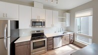 This thoughtfully designed single-floor unit offers a cohesive and modern aesthetic throughout. The kitchen is a standout feature, showcasing a galley-style layout with sleek white upper cabinetry and contrasting dark lower cabinets, white quartz countertops, a classic white tile backsplash, and a full suite of stainless steel appliances including an electric range, built-in microwave, dishwasher, and refrigerator. Track lighting overhead illuminates the workspace efficiently. Warm wood-look flooring flows consistently throughout the unit, connecting each area with a sense of continuity and warmth. The living area is open and filled with natural light from a large window that frames views of surrounding greenery, creating a bright and inviting atmosphere. The layout, as seen in the overhead dollhouse view, is well-organized with clearly defined zones for living, cooking, and sleeping, connected by a clean central corridor. The bathroom is finished with neutral grey walls, a full-size soaking tub with a curved shower rod and chrome fixtures, wood-look flooring, and a vanity with dark cabinetry. Overall, the unit presents a clean, contemporary design with practical finishes suited for comfortable everyday living.