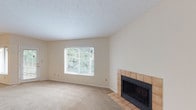 Located at 6112 Cedar Hill Ln in Florence, Kentucky, this single-floor home offers a comfortable and functional layout with a warm, neutral aesthetic throughout. The spacious living area is anchored by a tile-surround fireplace with a black insert, complemented by plush carpeting and a large window that frames lush, tree-lined outdoor views, filling the room with natural light. A textured ceiling adds a classic touch to the living space. Adjacent to the living area, a generous dining or secondary living space features a glass-panel door leading to an outdoor area, along with additional wide windows that continue to bring in ample daylight and greenery. The kitchen is thoughtfully designed with an open pass-through to the main living area, rich wood cabinetry, dark granite-style countertops, and a full suite of black appliances including a refrigerator, electric range, and built-in microwave. Tile flooring in the kitchen provides a practical contrast to the carpeted living spaces. A pendant light fixture adds a finishing touch to the dining area. The overall ambiance of the home is bright, open, and inviting, with a cohesive neutral color palette that creates a versatile canvas throughout.