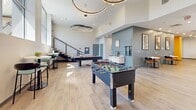 This expansive amenity lounge offers a thoughtfully designed, open-plan layout that seamlessly blends entertainment, socializing, and relaxation across a generously sized single floor. The space is anchored by light-toned hardwood flooring throughout, lending warmth and continuity to each distinct zone.

The games area is a standout feature, outfitted with a foosball table, a billiards table, and a shuffleboard table, all situated beneath striking circular pendant chandeliers that add a sculptural, upscale touch. High ceilings in this section amplify the sense of openness and energy.

Adjacent to the games area, a comfortable lounge zone features plush seating arranged around a wall-mounted flat-screen TV, framed by a textured accent wall. Floor-to-ceiling windows flood this area with natural light while offering impressive city skyline views.

The social dining area includes a built-in bar counter with bar-height seating and compact café-style tables with colorful accent chairs, creating an inviting space for casual gatherings. Vibrant pops of orange and yellow on structural columns add a lively, contemporary energy.

A kitchenette with dark countertops and custom cabinetry featuring a hexagonal tile accent wall provides practical functionality. Curated artwork adorns the walls throughout, contributing to the lounge's modern, urban aesthetic.