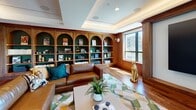 This thoughtfully designed single-floor space exudes refined elegance through a cohesive blend of warm materials and architectural detail. Rich walnut wood paneling lines the walls throughout, creating a warm and cohesive aesthetic that is both inviting and distinguished. A standout feature is the floor-to-ceiling built-in bookcase, showcasing a series of arched alcoves set against deep hunter green backdrops, providing striking contrast and a classic library-inspired atmosphere. The tray ceiling, accented with recessed and ambient lighting, adds architectural depth and a sense of elevated design. Hardwood flooring in a warm tone runs throughout, complementing the wood-paneled walls seamlessly. Large windows on multiple walls allow generous natural light to fill the room and offer views of the surrounding outdoor environment. The main seating area centers around a generous leather sectional sofa paired with a geometric-patterned area rug in muted greens and yellows, adding a contemporary touch to the otherwise traditional setting. Steel-framed glass sliding doors provide elegant visual connectivity to adjacent areas while maintaining a sense of openness. Decorative accents, including a sculptural gold feline figure and curated shelf displays, round out the space with personality and style.