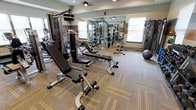 This fitness center offers a comprehensive workout environment designed to support a range of exercise routines. The space is outfitted with an array of strength training equipment, including multi-station cable machines, adjustable weight benches, and free weights, providing users with versatile options for resistance training. A stability ball and additional accessories are also available for functional fitness exercises.

The room features neutral-toned carpet with subtle striped accents, contributing to a clean and organized atmosphere. Large windows along the walls allow natural light to fill the space, creating a bright and energizing environment. Ceiling fans are mounted overhead, promoting air circulation throughout the workout area.

A full-length wall mirror spans one side of the room, visually expanding the space and allowing users to monitor their form during exercise. The walls are accented with horizontal color-block stripes in muted tones, adding a modern design element. The layout is thoughtfully arranged to allow comfortable movement between stations, ensuring an accessible and functional fitness experience.