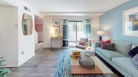 This two-story model unit is thoughtfully designed to maximize functionality across both levels, connected by a staircase that serves as a central architectural element. The main level features a welcoming living room finished with wood-look flooring, where a bold accent wall in soft blue anchors the space and a sliding glass door fills the room with natural light while providing outdoor access. The open layout allows the living area to flow comfortably, offering ample room for seating and entertaining.

The kitchen follows a galley-style configuration, lined on both sides with white cabinetry and white countertops that create a clean, bright aesthetic. White appliances, including a range, microwave, and dishwasher, are neatly integrated throughout.

Upstairs, the bedrooms are carpeted for comfort and feature windows that allow natural light to filter in. Each bedroom includes closet storage, with one room offering a walk-in style closet and another featuring a standard sliding-door closet with a dedicated rod and shelving system. One bedroom also connects directly to a bathroom area.

The bathroom is appointed with a white vanity featuring under-sink cabinetry, a single-basin sink, chrome fixtures, and a large mirror, all contributing to a clean and practical finish throughout the unit.