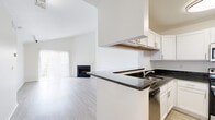 This single-floor residence offers a clean, modern aesthetic defined by crisp white walls and an open, airy layout. The living room is a standout space, showcasing soaring vaulted ceilings that amplify the sense of volume, light wood-tone flooring, and a fireplace as a focal point. A large sliding glass door with vertical blinds fills the room with natural light. The kitchen is thoughtfully designed with white shaker-style cabinetry, dark granite countertops, stainless steel appliances including a refrigerator and dishwasher, and a peninsula that connects to the living area through a pass-through opening, enhancing the open-concept flow. Light wood-tone flooring continues throughout the kitchen and main living areas. The bedroom features plush carpeting and bright white walls, providing a comfortable, neutral retreat. Two bathrooms serve the unit: one includes a walk-in shower with sliding glass doors and wood-look flooring, while the other features a soaking jetted tub with a shower and built-in shelving. A dedicated laundry area with a washer and overhead wire shelving adds everyday convenience to this well-appointed space.