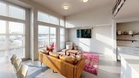 Located at 606 Burwell Street in Bremerton, Washington, this single-floor amenity space blends contemporary design with a rich nod to local history. The open-concept layout connects a comfortable lounge, a fully equipped kitchen, and a spacious dining area in a bright, cohesive environment.

The lounge area is anchored by a tan leather sofa, a vibrant magenta area rug, and a wall-mounted television. Floor-to-ceiling windows flood the space with natural light and offer sweeping views of the Bremerton waterfront and surrounding cityscape, creating an inviting atmosphere for relaxation or socializing.

The kitchen features white cabinetry, a stainless steel refrigerator, a gas range with a ventilation hood, subway tile backsplash, open wooden shelving, and a generous white quartz island with a sink — ideal for communal gatherings and events. A dramatic black-and-white photographic mural depicting vintage Bremerton street scenes and a historic panoramic cityscape wraps across the walls, serving as a distinctive artistic centerpiece that celebrates the area's heritage.

The dining area accommodates a large wooden table with seating for eight, set beneath soaring ceilings and surrounded by expansive windows that invite abundant daylight. French doors provide access to an outdoor area, further enhancing the connection to the vibrant Bremerton waterfront setting. Polished concrete flooring and clean white walls tie the entire space together with a modern, urban aesthetic.
