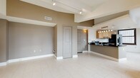 This well-appointed single-floor space showcases a thoughtfully designed open-concept layout that connects the living area to the kitchen with effortless flow. The interior is finished in warm taupe and crisp white tones, creating a clean, contemporary aesthetic throughout. Light-colored large-format tile flooring extends across the main living area, reflecting natural and artificial light to enhance the sense of openness. A ceiling-mounted track lighting system provides flexible, modern illumination and adds an industrial-chic touch to the space. The kitchen is outfitted with rich dark countertops that provide a striking contrast against warm wood-toned cabinetry, complemented by a multi-bulb pendant light fixture above. Appliances are neatly integrated into the cabinetry, contributing to a streamlined appearance. Structural ceiling details, including recessed soffits and defined transitions between spaces, add architectural interest and depth to the interior. Interior doors feature classic raised-panel designs in a muted gray finish, harmonizing with the neutral color palette. A wall sconce near the entryway adds a warm, welcoming accent. The overall ambiance is modern, clean, and versatile, making this a highly functional and visually cohesive living space.